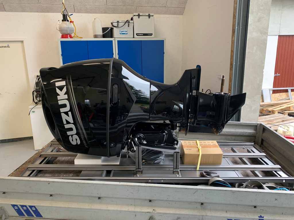 Ny sort Suzuki-påhængsmotor spændt fast på et stativ på en trailer inde i værkstedet, med blå værkstedsskabe og åben port bagved