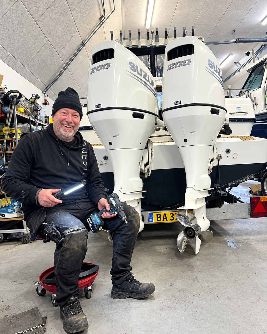 Rasmus Almbjørn sidder på en rullestol med arbejdslampe og skruemaskine i hænderne, foran to nye Suzuki 200 hk på agterspejlet
